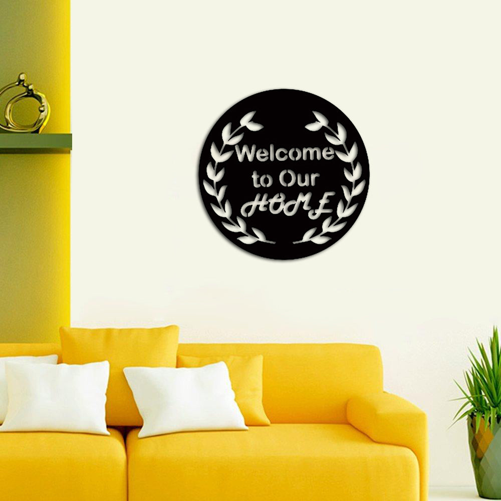  Black MDF circular wall art with the text "Welcome to Our Home" surrounded by a laurel wreath design, mounted on a light wall above a yellow sofa with white cushions.